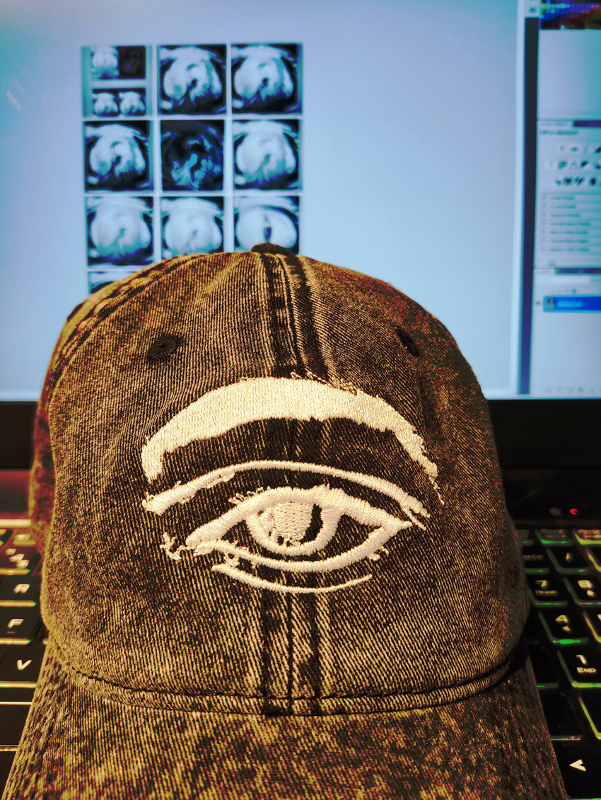 Kurt's 3rd Eye Denim Vintage Cotton Twill Cap by Emily Yap Design — minimalist white embroidered eye outline on a blue denim cap, resting on a studio desk in front of a laptop displaying surrealist graphite drawing reels