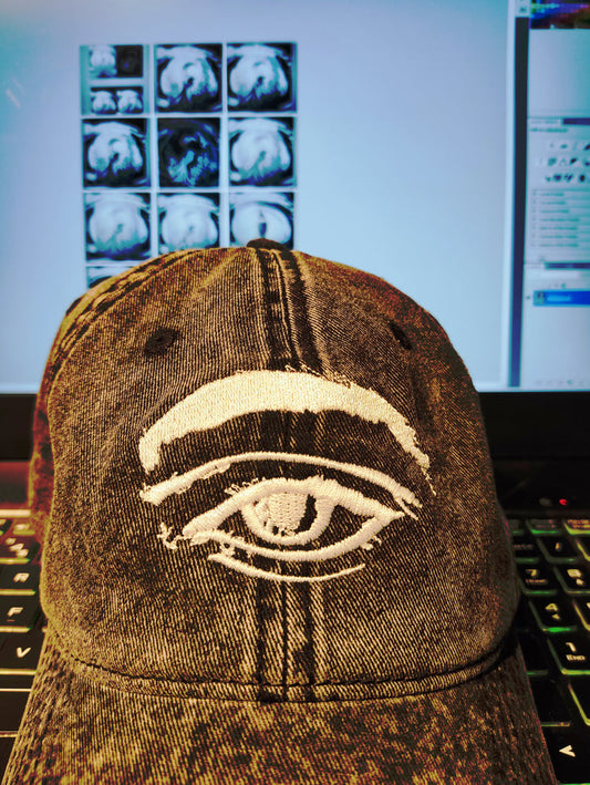 Kurt's 3rd Eye Denim Vintage Cotton Twill Cap by Emily Yap Design — minimalist white embroidered eye outline on a blue denim cap, resting on a studio desk in front of a laptop displaying surrealist graphite drawing reels