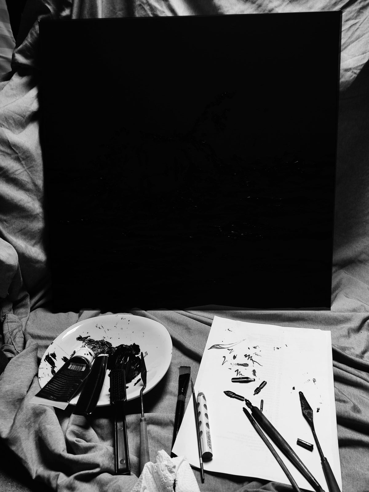 Black-and-white studio photograph by Emily Yap Design showing a black canvas, a paint-covered palette, brushes, palette knives, and marked paper